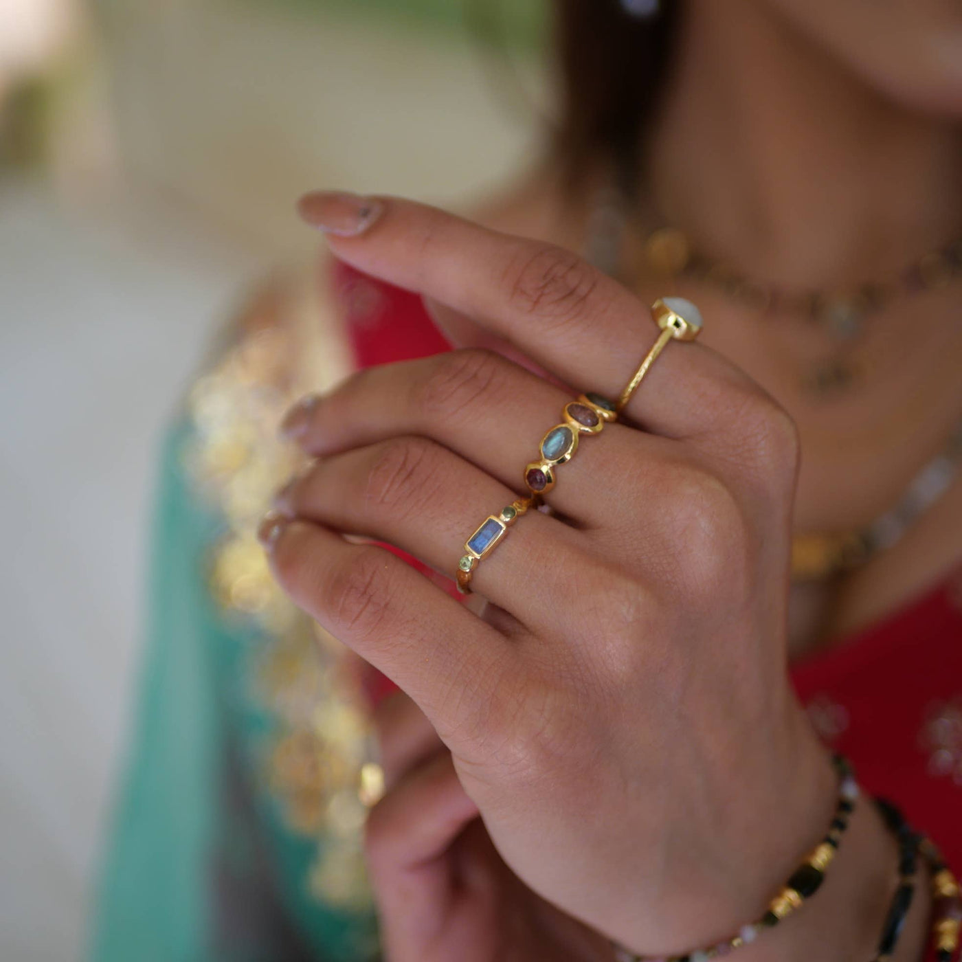 Nahaufnahme einer Hand mit drei goldenen Ringen, darunter ein Ring mit bunten Edelsteinen in Rosa, Blau und Grün, getragen vor einem farbenfrohen Outfit in Rot, Grün und Gold.