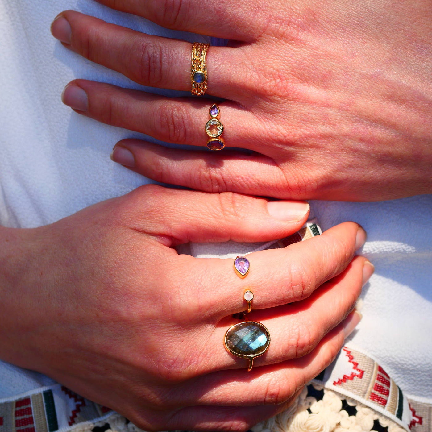 Hand mit verschiedenen Ringen, unter anderem "Paula" Goldring mit gehämmertem Effekt, besetzt mit Amethyst und Regenbogen-Mondstein, auf hellem Hintergrund.
