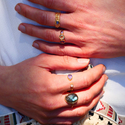 Hand mit verschiedenen Ringen, unter anderem "Paula" Goldring mit gehämmertem Effekt, besetzt mit Amethyst und Regenbogen-Mondstein, auf hellem Hintergrund.