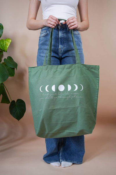 Frau hält eine große grüne Baumwolltasche mit Mondphasen-Print und Spruch „And just like the moon, you shall go through phases“ vor dem Körper – stehend in Jeans.