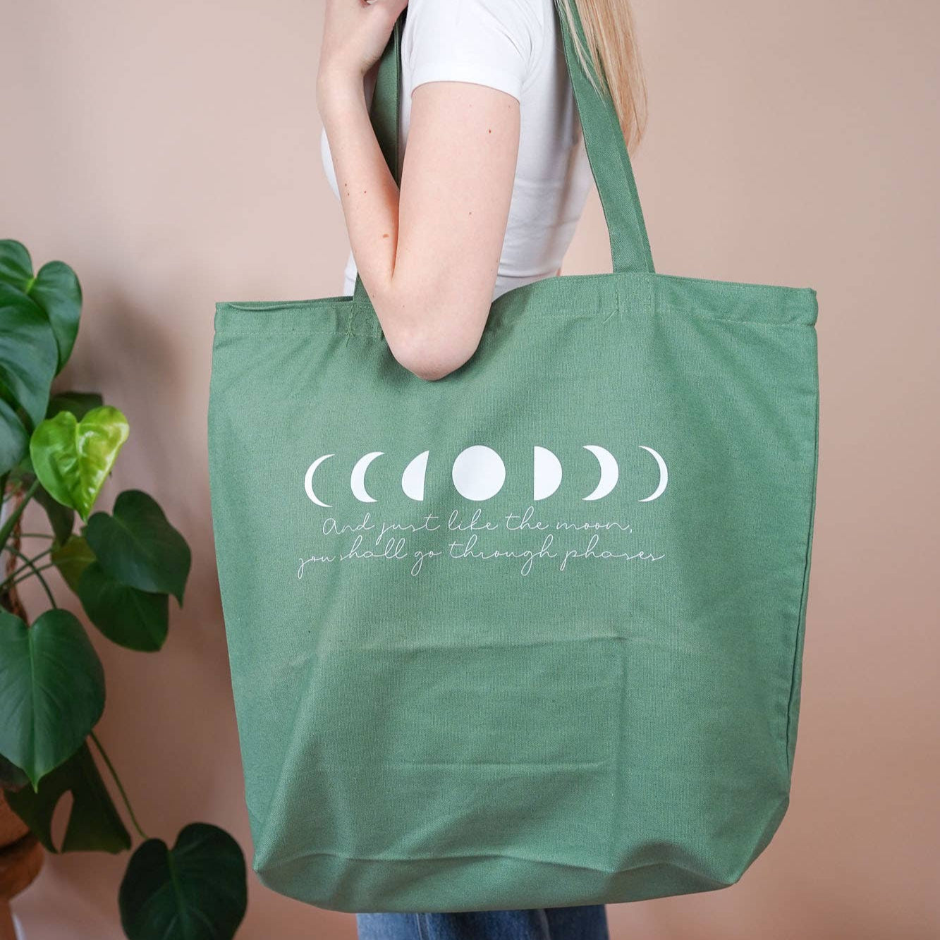 Grüne Baumwolltasche mit Mondphasen-Print und dem Spruch „And just like the moon, you shall go through phases“, getragen über der Schulter.